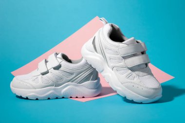 Makro kenarlı bir bakış açısı. Bir beyaz çocuk ayakkabısı ikinci spor ayakkabısının arkasında duruyor. Geometrik kare kağıt üzerinde pembe ve mavi arka plan.