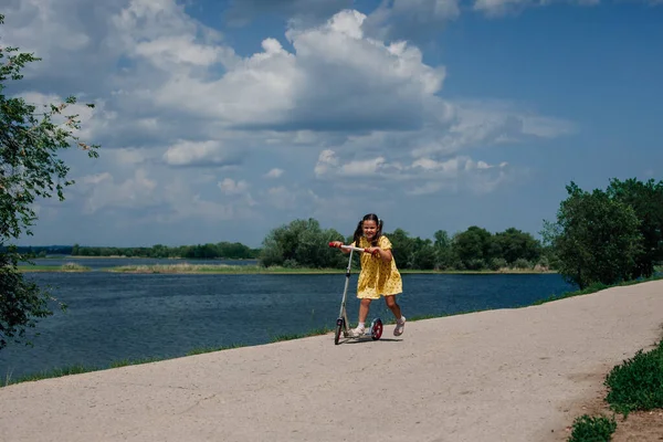 Stile di vita ritratto completo di un bambino ridente e felice che cavalca uno scooter in una bellissima foresta lungo un percorso lungo un fiume in una calda giornata di sole — Foto Stock