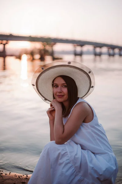 Picnic al atardecer en el puente sobre el río, mujer caucásica relajada en un vestido blanco y sombrero —  Fotos de Stock