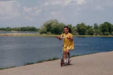Çocuklar spor aktiviteleri ve aile hafta sonları, bir çocuk hafta sonu parktaki göletin kenarındaki yolda scooter sürüyor.