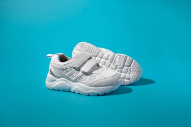 İki beyaz genç spor ayakkabısının yan görünüşü. Cırt cırtlı diğer bir ayakkabının tabanının önünde duran sert gölgeli mavi kağıt bir arka planda.