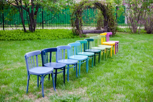 Una fila diagonal de sillas de madera de arco iris en un césped verde en el parque, preparándose para una fiesta familiar en un fin de semana al aire libre — Foto de Stock