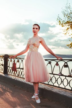 Genç bir bale dansçısı pembe bir elbise ve kurdeleli Çekler gün batımında sıcak bir yaz gününde nehir çitlerinin dökme demir parmaklıklarında duruyor.