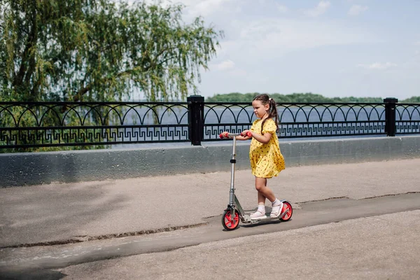 Uma menina em um vestido amarelo aprende a andar de scooter dado para seu aniversário ao longo do lago da cidade — Fotografia de Stock