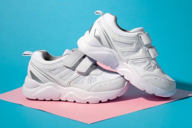 Geometrik kare bir kağıtta cırt cırt cırtlı iki beyaz spor ayakkabının yan görüntüsü. Pembe ve mavi arka planda sert gölgeler var. Bir spor ayakkabı diğerinin üzerinde duruyor.