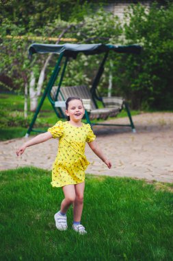 Gülen bir kız arka planda bir bahçe salıncağıyla yaz bahçesinde dans eder ve sıcak bir günün tadını çıkarır.