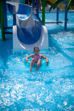 Su parkındaki su kaydırağından yüzen pembe mayo giymiş beş yaşındaki gülümseyen bir kız çocuğu. Aktif eğlence ve sertleşme.