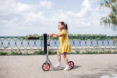 Mutlu, gülümseyen bir kız şehirde nehir kıyısında scooter sürüyor. Güneşli bir yaz gününde motosiklet sürmeyi öğreniyor.