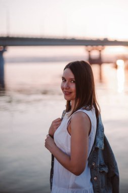 Kot gömlekli, beyaz elbiseli, zarif, gülümseyen bir kadının portresi pembe bir gün batımı ve nehir üzerinde bir köprü.