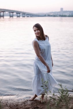 Beyaz bir yaz elbisesi içinde, gün batımında bir nehir ve arkasında bir köprü olan genç ve güzel bir kadın. Pastel yumuşak renklerde yaz günbatımı. Romantik randevu kavramı