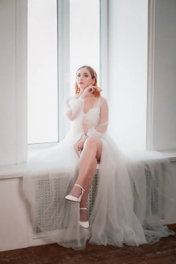 Beyaz şeffaf pelerinli, pencerenin kenarında oturan ve elini yüzüne dayayan masum bir kadının tam boy portresi.