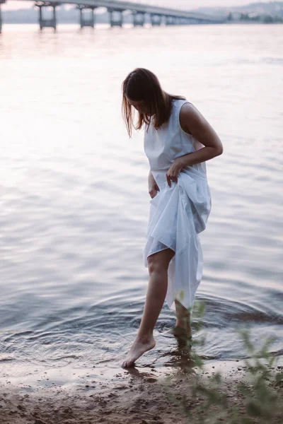 Eine schlanke junge Frau in einem weißen Sommerkleid, die den Saum ihres Kleides hebt und ihre Füße nach Sonnenuntergang im Wasser am Sandstrand des Flusses benetzt — Stockfoto