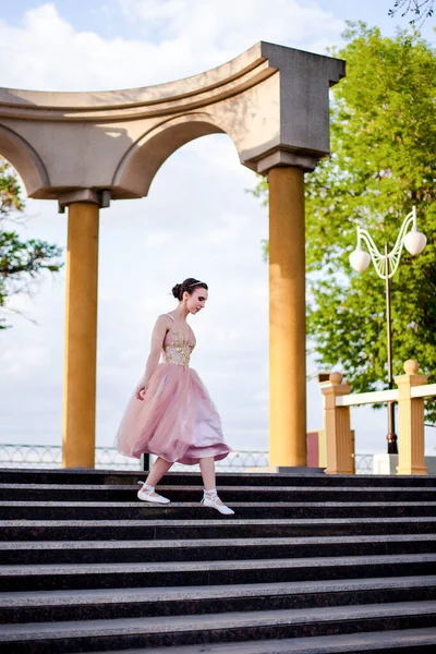 ピンクのシルクのドレスを着た細い女性のバレリーナとリボンのついた靴が優雅に夏の日没時に公園の列とアーチの階段を降ります — ストック写真