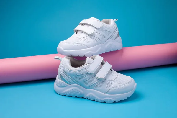 Side view balance of two white unisex sneakers on a pink long paper roll on a blue background, jeden sneaker stoi na drugim sneakerze — Zdjęcie stockowe