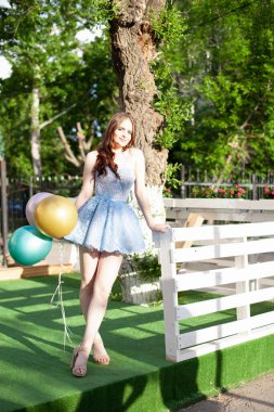 Neşeli, kısa mavi bir korse giymiş mutlu bir kadın. Parlak güneş ışığı altında beyaz ahşap tahtalardan bir çitle bir yazlık kafenin terasında duruyor.