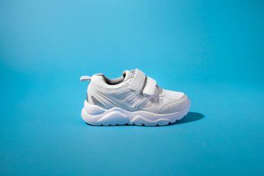 İzole edilmiş mavi bir arka planda, cırt cırt cırtlı ayakkabı bağlayıcıları olan yeni bir beyaz çocuk ayakkabısı.