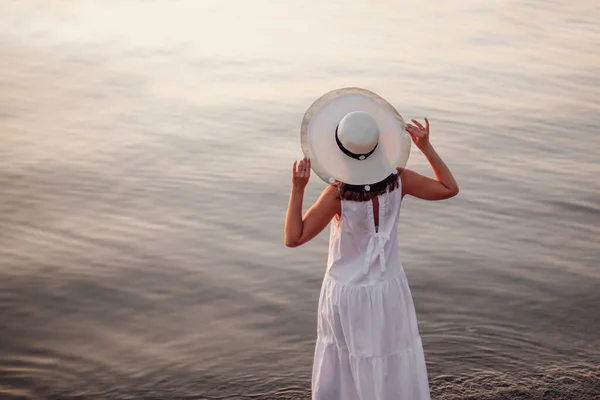 一个戴帽子的女人在海边日落时，一个戴着草帽、身穿白色衣服、背对着大海的女人的生活风格画像 — 图库照片