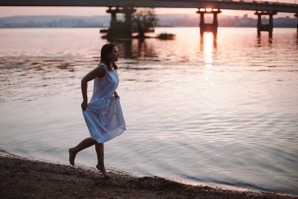 Mulheres a correr ao pôr-do-sol. Uma jovem encantadora em um vestido de verão branco correndo descalça na praia de areia do rio ao pôr do sol — Fotografia de Stock