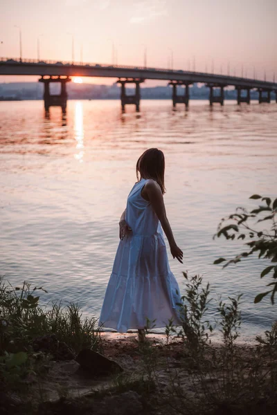 Die Silhouette einer Frau, die bei Sonnenuntergang tanzt. Lifestyle-Ganzkörperporträt einer tanzenden jungen Frau in einem langen weißen Sommerkleid bei Sonnenuntergang am Strand in der Nähe der Brücke über die Brücke. — Stockfoto