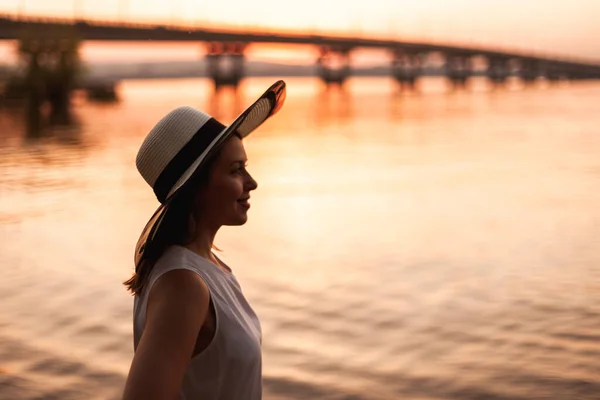 Una mujer en el puente sobre el río al atardecer. Retrato de perfil de una joven con sombrero de paja admirando la puesta de sol en la playa de un río con un puente en una noche de verano —  Fotos de Stock