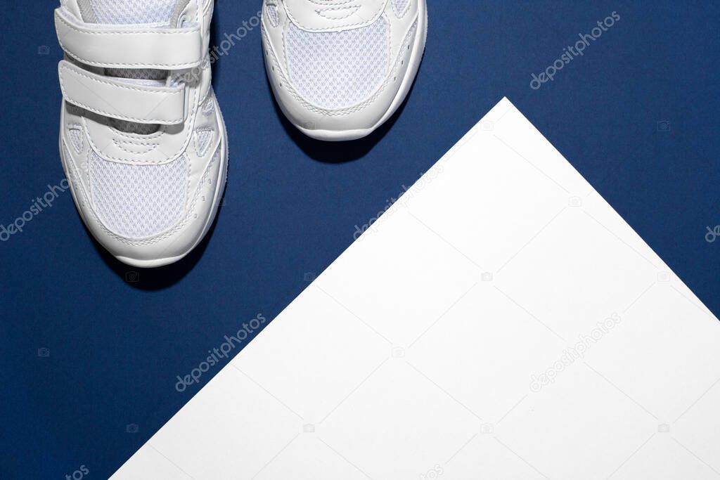 Photo of a pair of white childrens sports sneakers made of leather and sewn with fabric with a velcro fastener with shadows on a blue and white background with a place to copy text or design