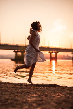 Gün batımında koşan kadınlar. Uzun beyaz elbiseli beyaz bir kadının portresi turuncu, parlak yaz günbatımında köprüsü olan bir nehrin sahilinde uzanıyor.