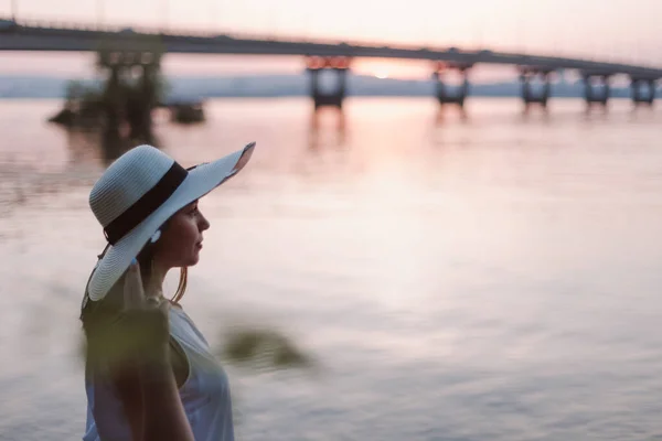 日落时在河岸上的女人日落时分，一个头戴草帽的白人妇女在河边的桥上与城市的轮廓一起放松沉思，这幅肖像是她的肖像 — 图库照片