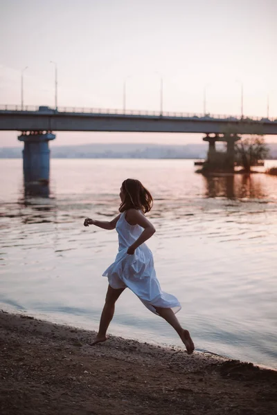 Eine Frau läuft am Ufer des Flusses entlang. Lifestyle-Porträt einer jungen Frau im weißen Kleid, die an einem heißen Sommertag bei Sonnenuntergang barfuß auf einem sandigen Flussufer mit einer Brücke läuft — Stockfoto