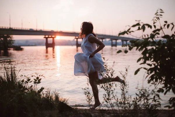 Mujeres corriendo al atardecer. Estilo de vida retrato de larga duración de una hermosa joven con un vestido blanco largo corriendo a lo largo de la orilla del río al atardecer —  Fotos de Stock