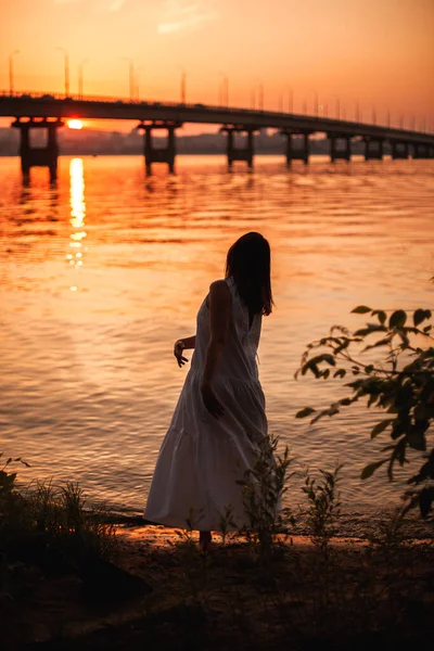日落时跳舞的女人的轮廓.在河边桥附近的海滩上，一位身材苗条的女人身穿白色的太阳衣，在橙色的夕阳下跳舞，她的生活风格是全长的. — 图库照片
