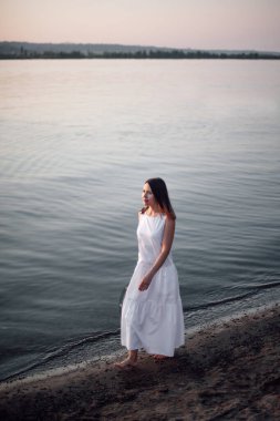 Sahilde bir kadın yürüyor. Güzel beyaz elbiseli ve yalınayak deniz kıyısında yürüyen, rahatlayan ve ruhsal ve zihinsel sağlığıyla ilgilenen genç bir kadının tam boy portresi.