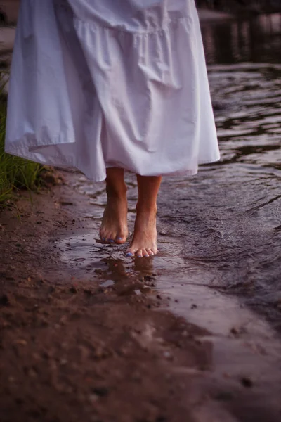 Frauenfüße im Fluss. Nahaufnahme der Füße einer Frau in einer langen Sommersonne, die an einem Sommerabend am Ufer des Sees spaziert — Stockfoto