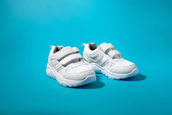 Vista frontal de dos zapatos deportivos blancos para niños con cierres de velcro para facilitar el calzado sobre un fondo de papel azul con luz solar dura —  Fotos de Stock