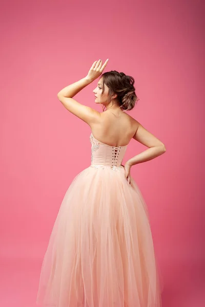 A young woman stands with her back against an isolated background. A beautiful woman model in a beige ball gown and evening hairstyle with bent arms poses on a pink background