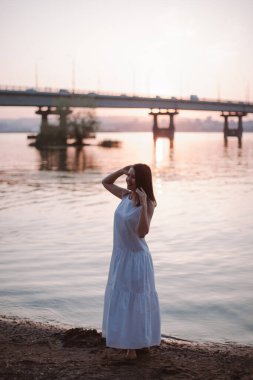 Gün batımında poz veren kadınlar. Gün batımında sahilde poz veren güzel bir genç kadının uzun bir portresi. Nehir üzerindeki köprüyü ve şehrin siluetini görüyor..