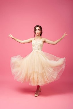 Genç bir kadın uzun bej bir balo elbisesi içinde kolsuz dans ediyor. Süslü korsesi ve bej ayakkabıları olan gür elbiseli esmer bir kadın izole bir arka planda dönüyor.