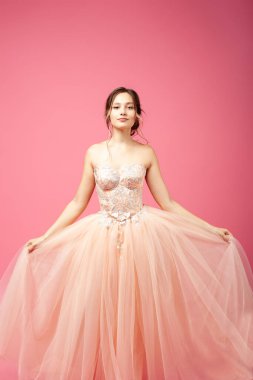 Güzel bir kadın yüzü ile ayakta duruyor ve ellerinde yemyeşil bir organza etek tutuyor. Kabarık şeftali rengi balo elbisesi ve akşam makyajı giymiş genç bir kadın pembe izole bir arka planda poz veriyor.