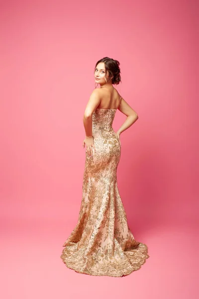 A young woman in a sparkling dress stands with her back to an isolated background. A beautiful young woman in a long dress and evening hairstyle poses on a pink background
