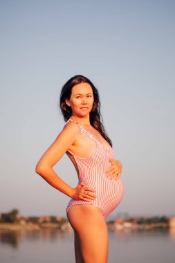 Mayolu hamile bir kadın, genç ve hamile bir kadının portresi. Yazın gün batımında plajda pembe mayo giymiş.