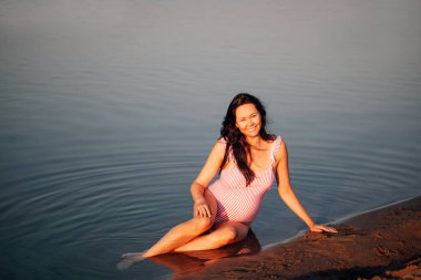 Kumsalda hamile bir kadın, huzurlu, rahat, pembe mayo giymiş hamile bir kadın yaz gezisinde denizin kumlu sahilinde oturuyor.