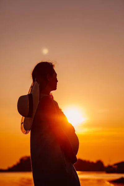 The silhouette of a pregnant woman at sunset. The silhouette of a young woman in a straw hat holding her pregnant belly with her hands on the sea with the setting orange vivid sun — Φωτογραφία Αρχείου