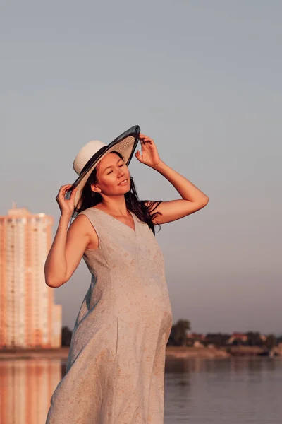 Una mujer embarazada al atardecer. Una mujer embarazada tranquila y relajada sostiene el borde de un sombrero de paja con sus manos y disfruta de la naturaleza y la armonía interior —  Fotos de Stock