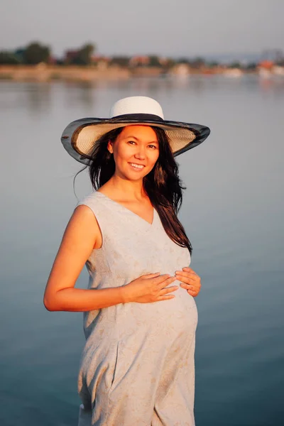 Una mujer embarazada al atardecer. Una mujer embarazada feliz y sonriente con sombrero de paja y vestido posa al atardecer junto al lago en una cálida noche de verano. —  Fotos de Stock