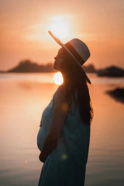 一个身披草帽、头戴草帽的亚洲女人的侧影。夏天，在橙色温暖的夕阳西下，她在海边抱着怀孕的肚子 — 图库照片