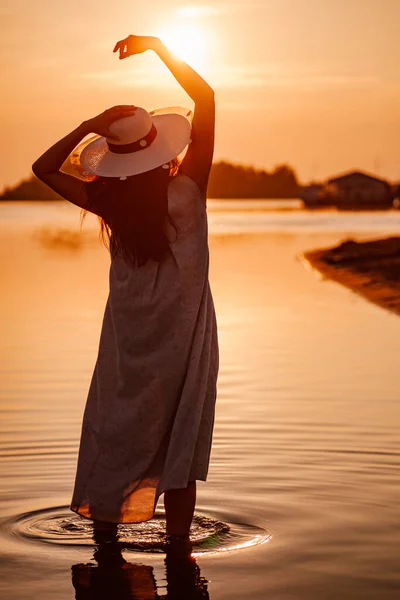 A silhueta de uma mulher ao pôr do sol. Uma silhueta de uma visão traseira de uma jovem mulher em um chapéu de palha com as mãos levantadas acima de sua cabeça em uma praia do mar em um pôr-do-sol laranja — Fotografia de Stock