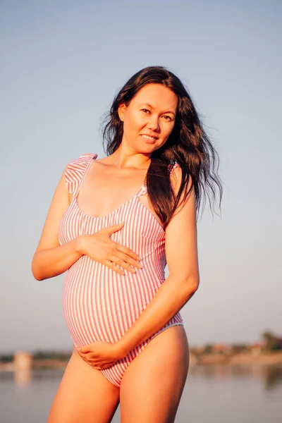 Ciężarna kobieta w kostiumie kąpielowym, uśmiechnięta, seksowna azjatka w ciąży brunetka w różowym stroju kąpielowym trzyma brzuch ciężarnej z rękami na plaży o zachodzie słońca — Zdjęcie stockowe