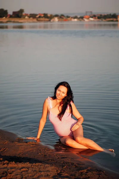 Mujer embarazada en la playa, sonriente, hermosa mujer embarazada en traje de baño rosa posando en la playa de arena del río en el pueblo —  Fotos de Stock