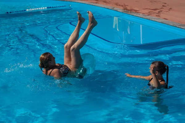 Abuela y nieta en la piscina. La abuela cae boca abajo de un círculo inflable en el agua de la piscina, en busca de equilibrio. Felices y divertidos momentos familiares — Foto de Stock