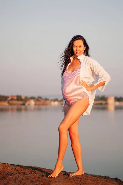 A pregnant woman on the beach, A beautiful, sexy pregnant woman in a pink swimsuit and a white shirt poses with her hands on her waist by the water at sunset on a summer vacation — Stock Photo, Image