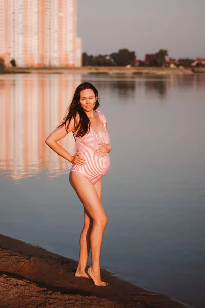 Femme enceinte en maillot de bain, portrait complet d'une femme brune asiatique enceinte posant en maillot de bain rose tenant la main sur un ventre enceinte pendant les vacances d'été — Photo